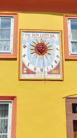 Detail malovaných slunečních hodin s červeným sluncem a latinským nápisem na žluté fasádě