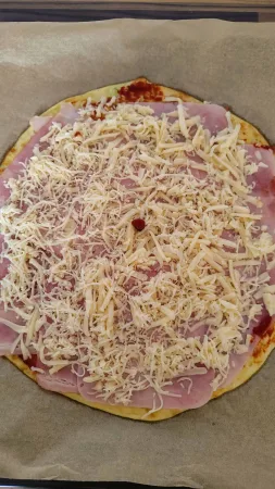 Cuketová pizza obložená plátky šunky a strouhaným sýrem, pohled shora před dopečením