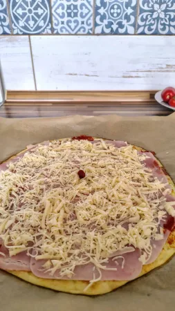 Předpečený korpus cuketové pizzy s rajčatovou omáčkou, šunkou a strouhaným sýrem před dopečením