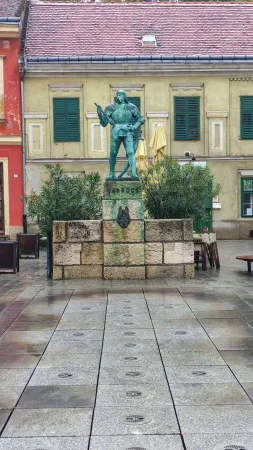 Bronzová socha muže na kamenném podstavci uprostřed dlážděného historického náměstí