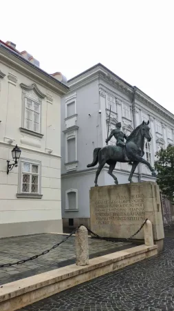 Bronzová jezdecká socha bojovníka s kopím na koni před historickými domy náměstí