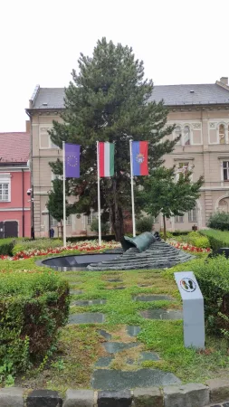 Malý parčík s evropskou a maďarskou vlajkou, stromem a pomníkem u dlážděných cestiček