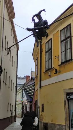 Pohled na sochu šaška visícího nad úzkou uličkou s kolemjdoucími