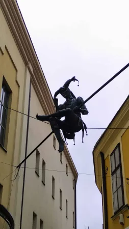 Bronzová socha šaška zavěšená na drátě nad uličkou mezi domy