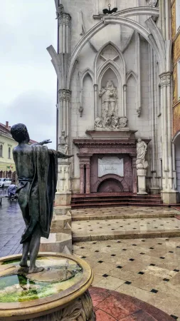 Bronzová socha muže u kamenné fontány před gotickými zříceninami v parku Romkert