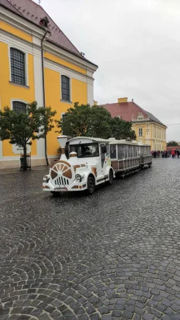 Bílý turistický vláček s vagóny projíždějící dlážděným náměstím kolem žlutého kostela