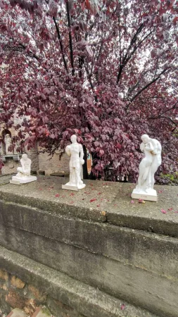 Tři bílé sochy na zídce pod fialovým stromem v zahradě