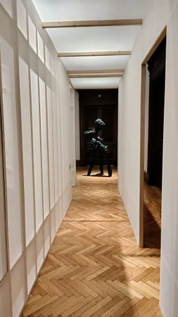 Bílá chodba galerie s dřevěnou podlahou a bronzovou sochou v pozadí