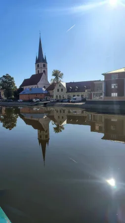 Kirchturm und umliegende Häuser, die sich im Gegenlicht der Sonne auf der Sázava spiegeln