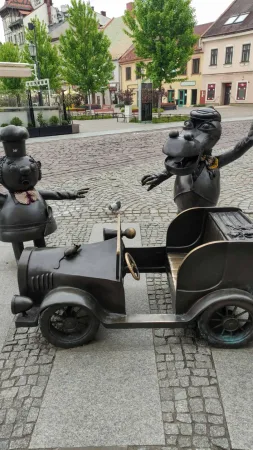 Bronzová socha starého automobilu s postavičkami kuchaře Bartolini a draka na náměstí.