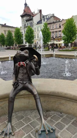 Bronzová socha žáby s deštníkem a kloboukem sedící na okraji kašny na náměstí.