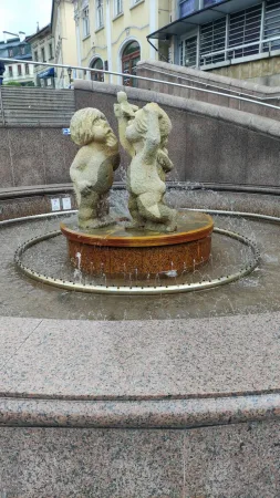 Kamenná fontána se sochou dvou dětí uprostřed tryskající vody na náměstí.