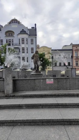 Kamenná fontána se sochou uprostřed obklopená historickými domy na náměstí.