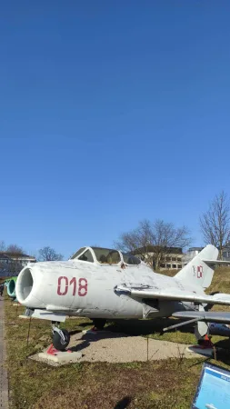Bílý stíhací letoun MiG s označením 018 vystavený venku na travnaté ploše.