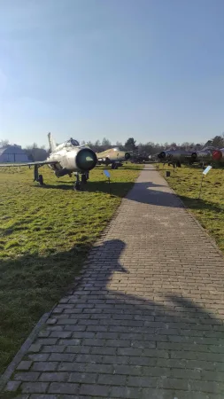 Dlážděný chodník vedoucí k vystavenému stíhacímu letounu MiG pod modrou oblohou.