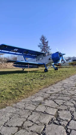 Modrobílý dvouplošník Antonov An-2 vystavený na trávě před muzeem letectví.