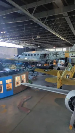 Expozice hangáru s kamuflovaným letounem, žlutým stíhacím letadlem a maketou modrého domku.