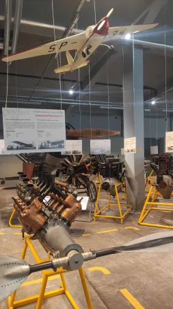 Zavěšený model historického letadla nad expozicí starých leteckých motorů.
