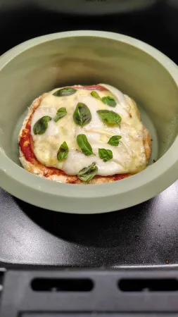 Upečená špaldová pizza s rozpuštěným sýrem a čerstvými lístky bazalky v zelené silikonové formě