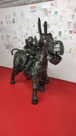 Kovová socha oslíka s malým jezdcem před nástěnkou dětských kreseb.