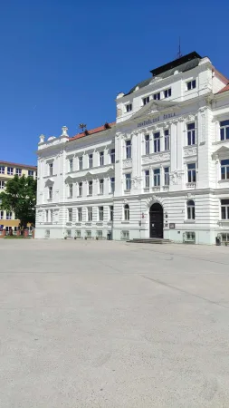 Bíle zdobená budova školy s vchodem a prázdným dlážděným prostranstvím před ní pod modrou oblohou