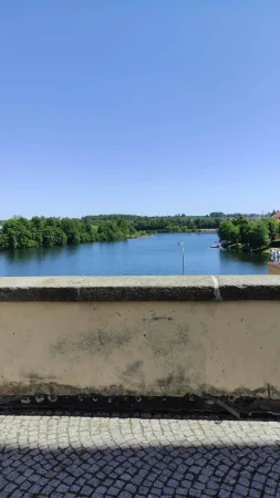 Výhled na modrou hladinu vodní nádrže Jordán obklopenou zelení z kamenné zídky nad cestou
