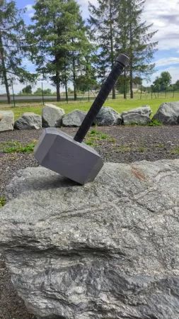 Replika Thorova kladiva Mjölnir zaražená do balvanu obklopeného jehličnatými stromy