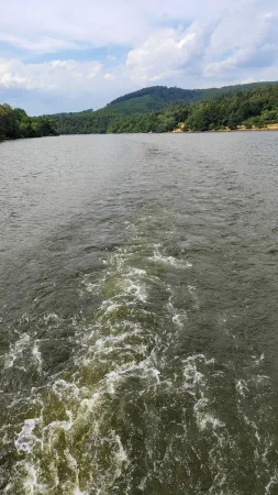 Pěnivá brázda za lodí s výhledem na zalesněné kopce v pozadí.
