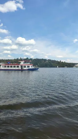 Výletní loď pluje po Brněnské přehradě obklopená zalesněnými kopci a modrou oblohou.