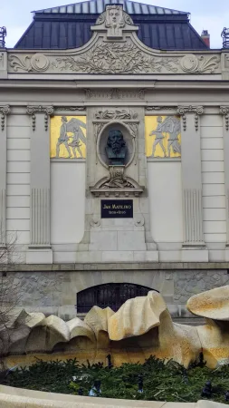 Bronzová busta malíře Jana Matejka se zlatými mozaikami na fasádě budovy v Krakově, před ní kamenná kašna