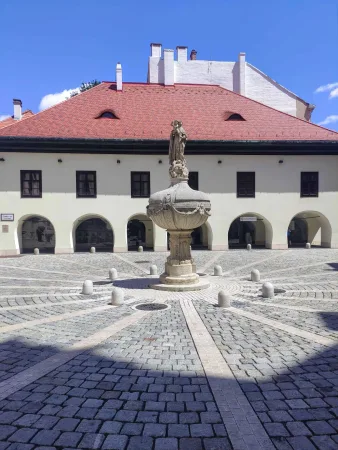 Kőudvar szökőkúttal és szoborral árkádos történelmi épület előtt Sopronban