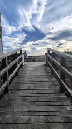 Dřevěná lávka vede k sedící postavě sledující západ slunce u hradních zdí.