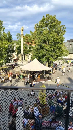 Letní koncert na historickém náměstí s morovým sloupem a davem diváků.