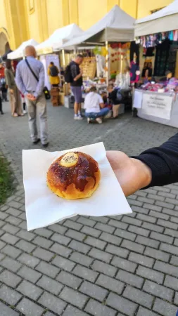 Ruka drží čerstvě upečenou sladkou pochoutku na tržnici před žlutou budovou.