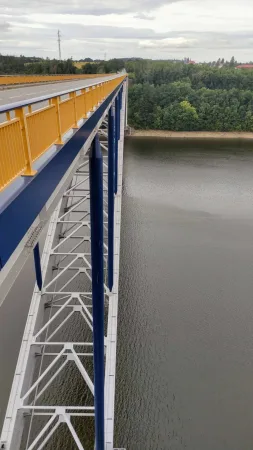 Detail žluto-modré ocelové konstrukce Žďákovského mostu nad zalesněným údolím Orlické přehrady